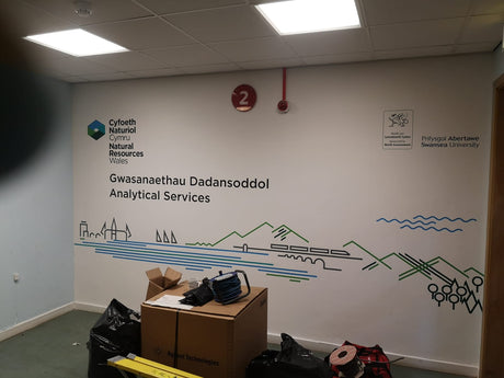 Room with a wall featuring a logo and text, with boxes and equipment on the floor.