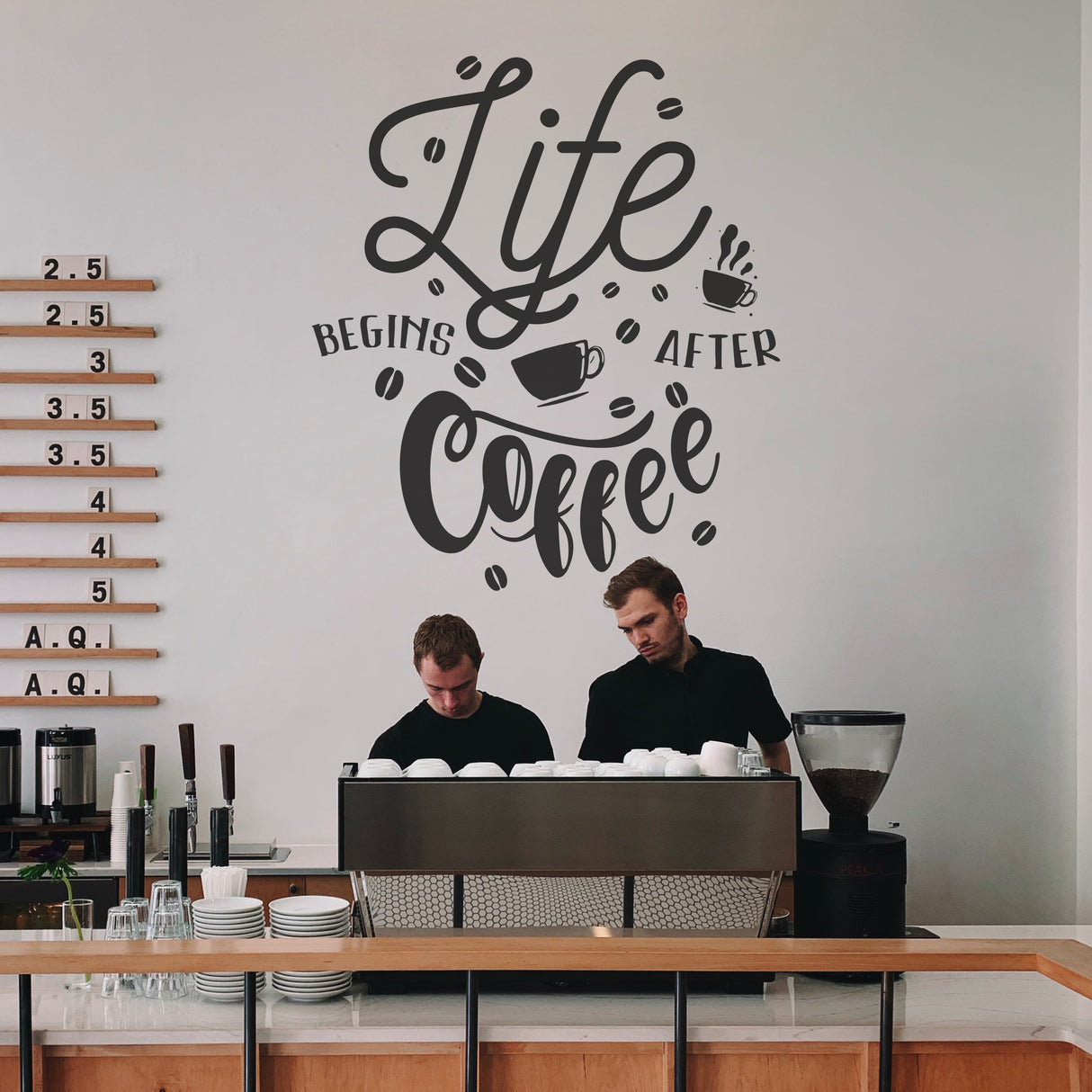 Two baristas working behind a counter with a 'Life Begins After Coffee' wall decal.